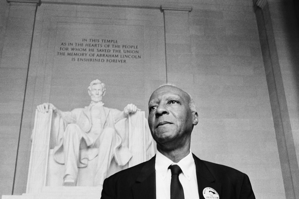 a philip randolph stands in front of a statue of abraham lincoln memorial, randolph wears a suit and tie with a button on his jacket