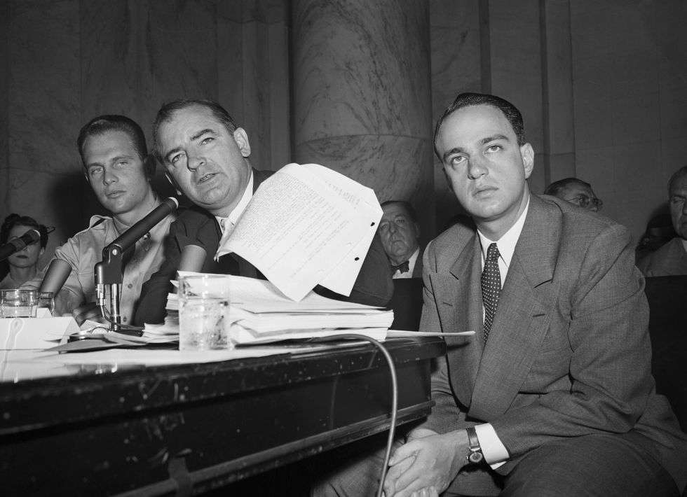 joseph mccarthy flipping through papers as he speaks into a microphone while roy cohn sits in a chair next to him with his hands calsped