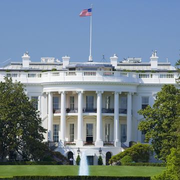 view of the white house on a bright sunny day, a fountain and green lawn are visible before the white building which has an american flag waving in the wind on top