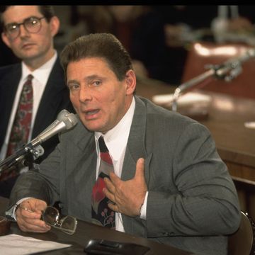 salvatore "sammy" gravano speaks into a microphone as he places one hand on his chest and looks to the right, he wears a gray suit jacket, white collared shirt and multicolor tie