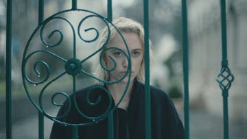 maika monroe in watcher, a young woman with blond hair standing behind some gates looking concerned