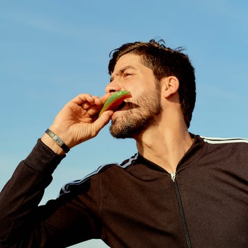 Man enjoying a slice of watermelon against a clear blue sky Person wearing a black athletic jacket outdoors holding an object to their mouth