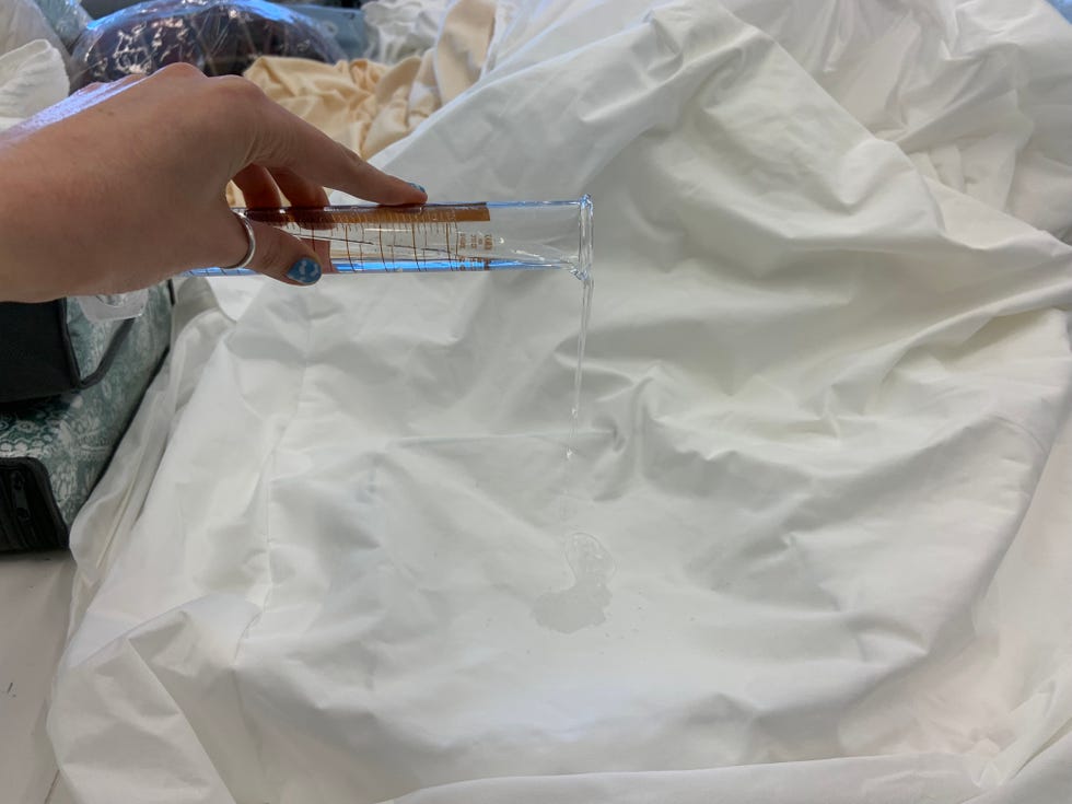 a lab analyst is pouring water onto a mattress protector from a beaker to analyze water repellency for good housekeeping's best waterproof mattress protectors