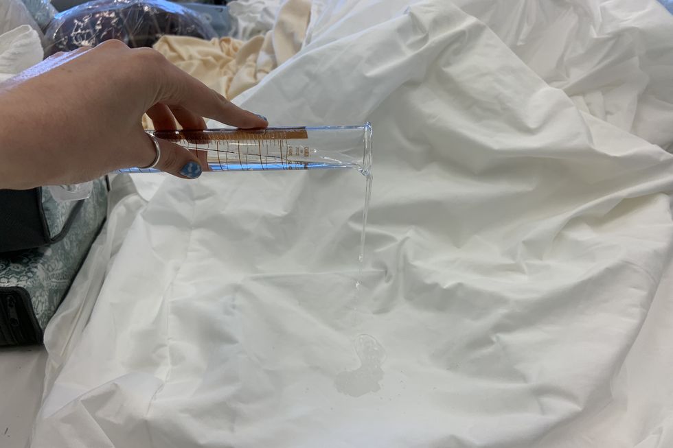 a lab analyst is pouring water onto a mattress protector from a beaker to analyze water repellency for good housekeeping's best waterproof mattress protectors