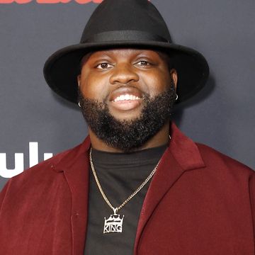 wavyy jonez, a man stands smiling at the camera, he has a black beard, he wears a black top and trousers with red shirt and black hat