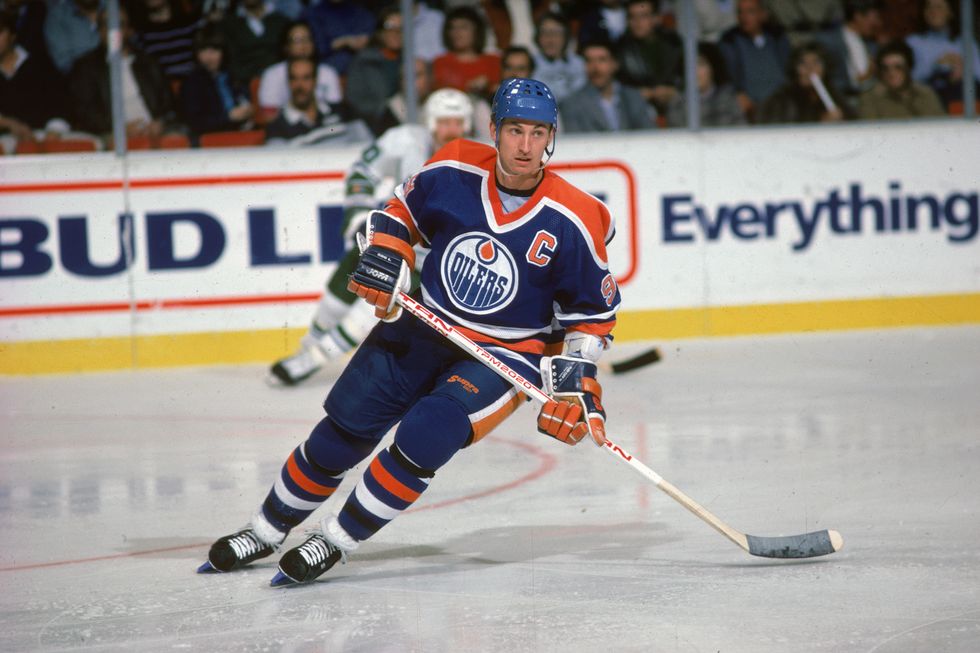 wayne gretzky skates on ice while wearing a blue and orange hockey uniform and equipment, he holds a hockey stick