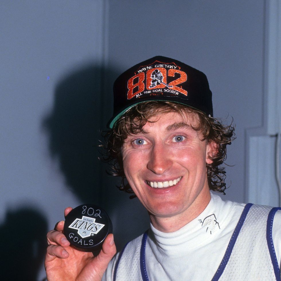 wayne gretzky smiles at the camera and holds up a hockey puck, he wears a black baseball cap with the number 802 in orange