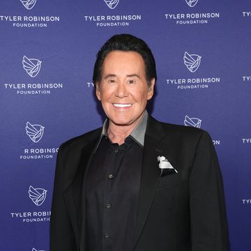 wayne newton smiles at the camera and stands in front of a blue background with a foundation logo on it, newton wears a black suit with a black collared shirt and pocket square