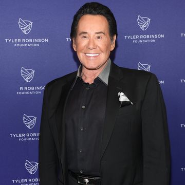 wayne newton smiles at the camera and stands in front of a blue background with a foundation logo on it, newton wears a black suit with a black collared shirt and pocket square