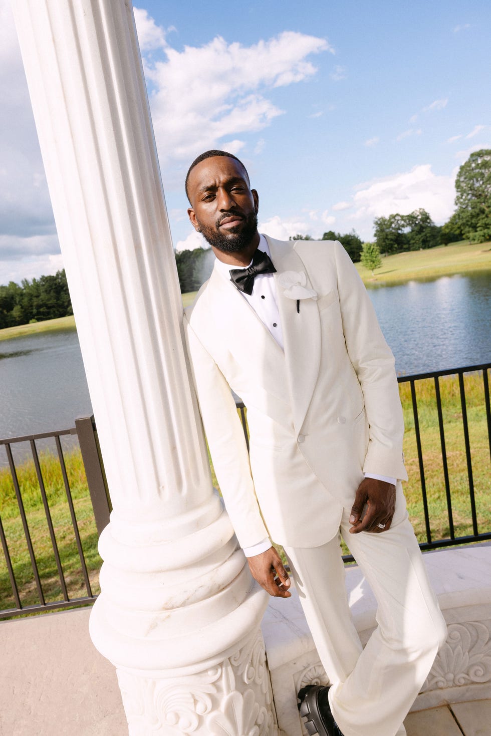 a man wearing a white suit leans on a column in front of a pond