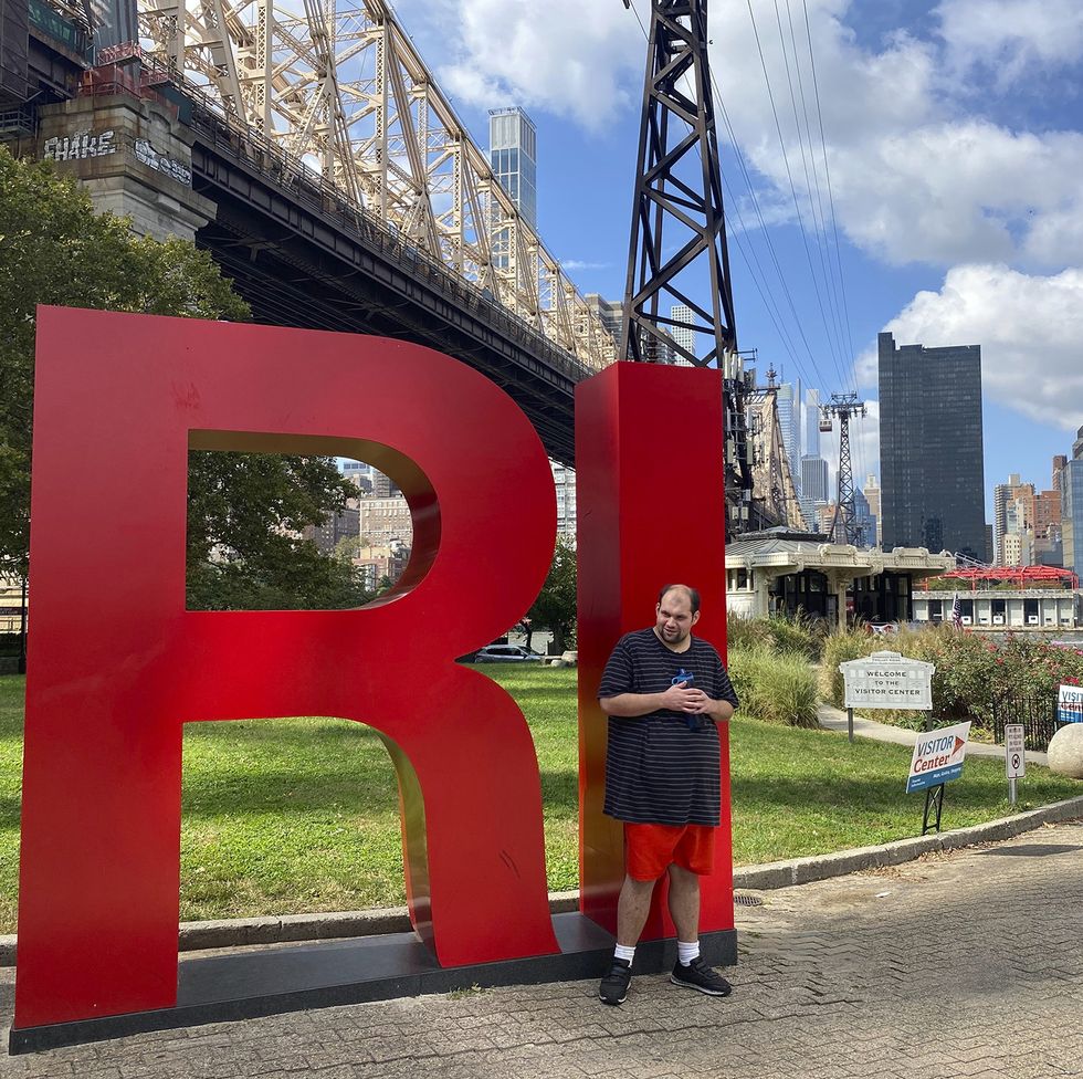 tony poses in front of a sculpture that says ri with a park and city skyline behind him