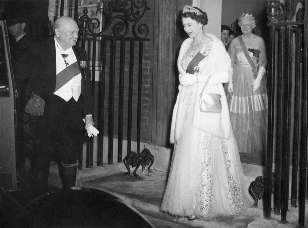 winston churchill holds a car door open and watches queen elizabeth walk toward it, he wears a tuxedo with a sash, she wears a gown with a fur stole, sash, and crown