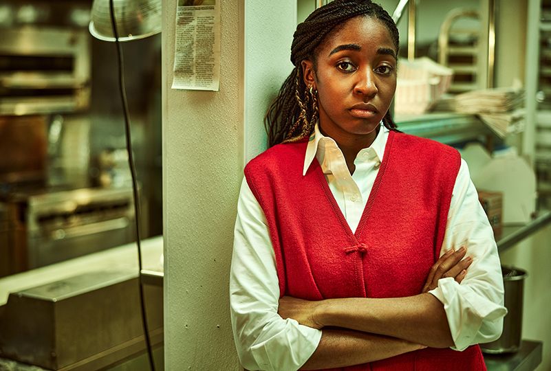 ayo edebiri in character as sydney on the bear stands in a kitchen with her arms crossed, she wears a white collared shirt with a red sweater vest on top