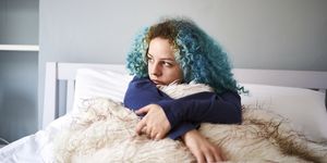 young woman with dyed blue hair looking sad whilst sat up in bed and gazing off into the distance