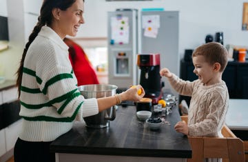 a boy in a beige sweater stands on a learning tower in the kitchen to help his mom who is in a white and green sweater and holding an orange