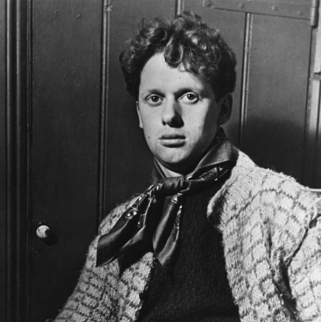 dylan thomas looks at the camera as he sits in a chair, he wears a sweater, scarf and dark top
