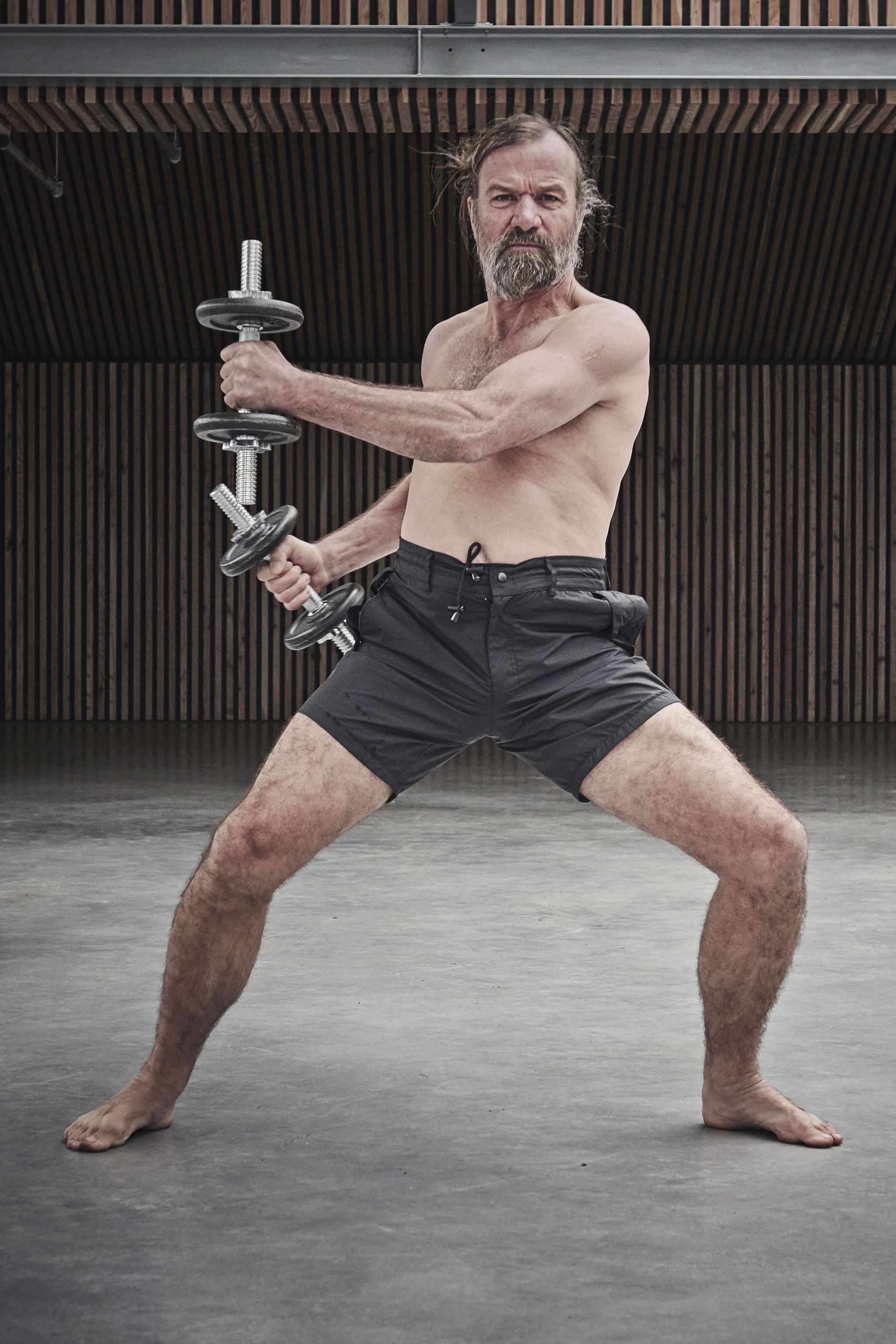 topless male middle aged bearded man standing barefoot holding a dumbell in each hand
