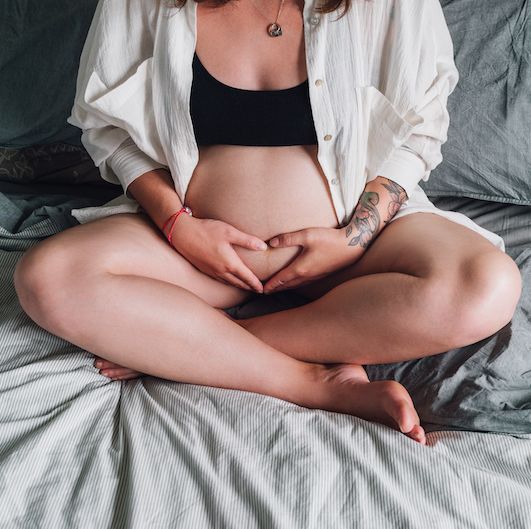 young pregnant woman with tattooed hand sitting cross legged touching her belly on a bed in soft light
