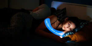 a woman lying in bed texting while her partner sleeps next to her