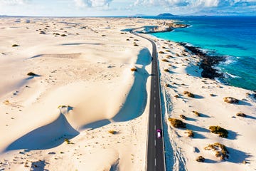 car with surfboard traveling on empty road in between sand dunes and ocean, corralejo , fuerteventura, canary islands, spain