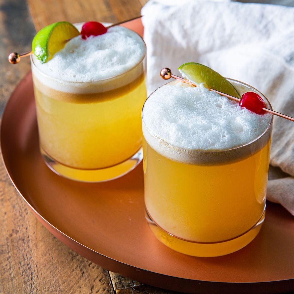 two whiskey sours garnished with maraschino cherries and lime on a copper tray