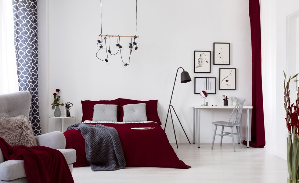 White and burgundy contemporary bedroom interior with a bed dressed in cotton linen and pillows. Framed art above small desk. Real photo.