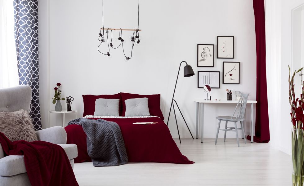 White and burgundy contemporary bedroom interior with a bed dressed in cotton linen and pillows. Framed art above small desk. Real photo.