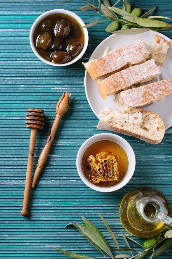 White bowls with sweet green olives in honey sauce and honeycombs, served with wood dipper, bottle of olive oil, sliced bread and olive branch over turquoise texture background. Top view