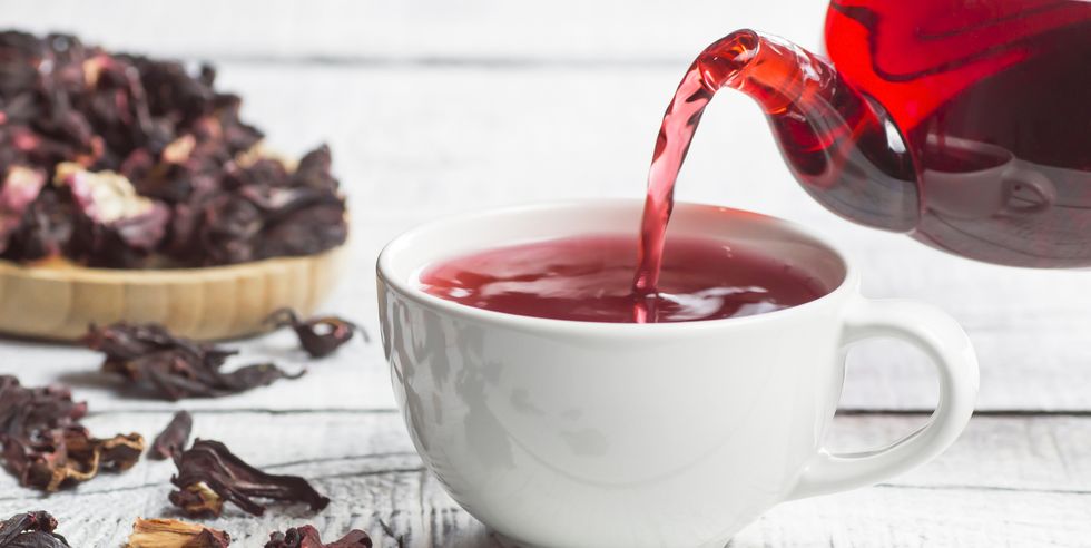 white cup of healthy hibiscus tea pouring from the teapot with dried hibiscus flowers on white wooden background, winter hot drink concept for cold and flu