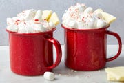 white hot chocolate in red mugs topped with whipped cream, crushed peppermint candies, white chocolate pieces, and mini marshmallows
