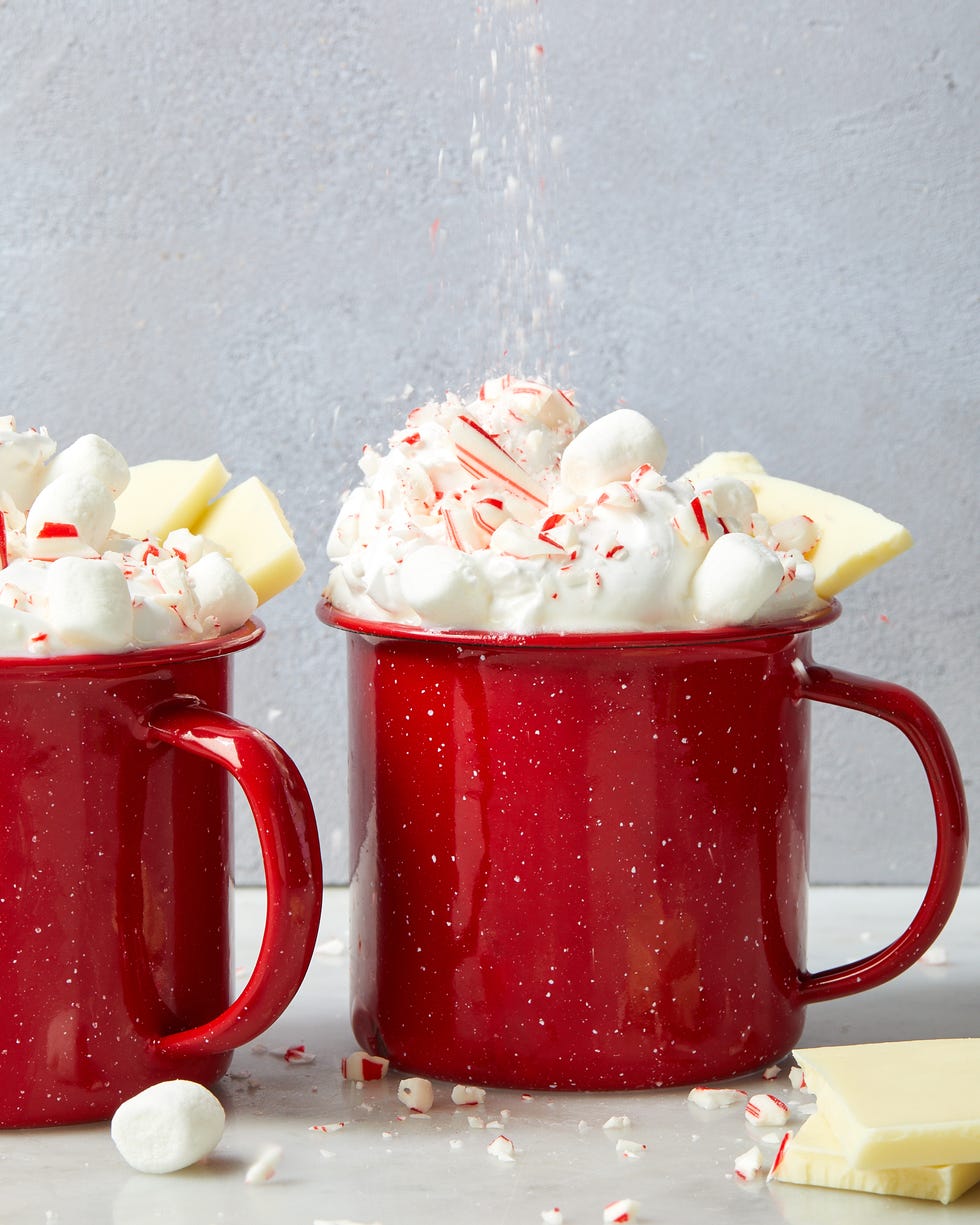 white hot chocolate in red mugs topped with whipped cream, crushed peppermint candies, white chocolate pieces, and mini marshmallows