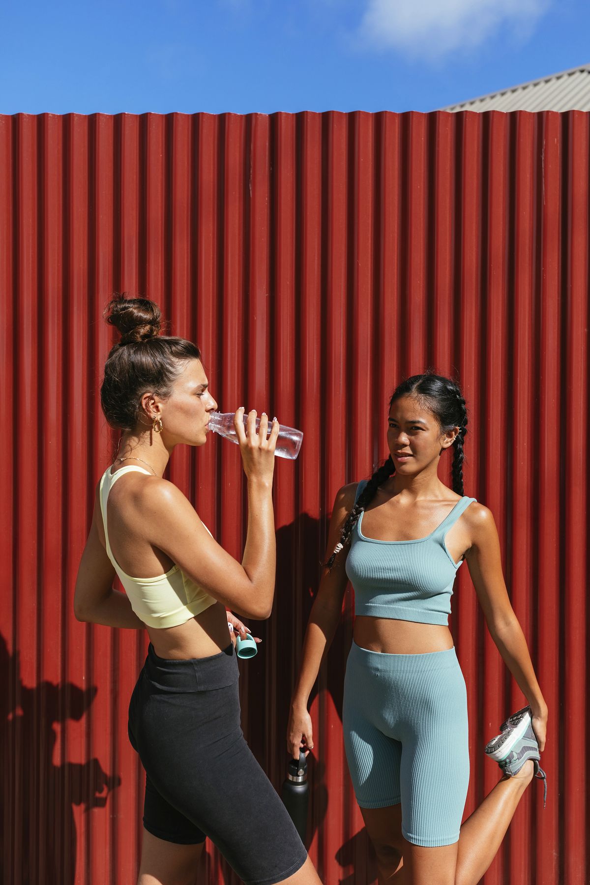 two female friends wearing sportswear, standing in front of the red wall and drinking water