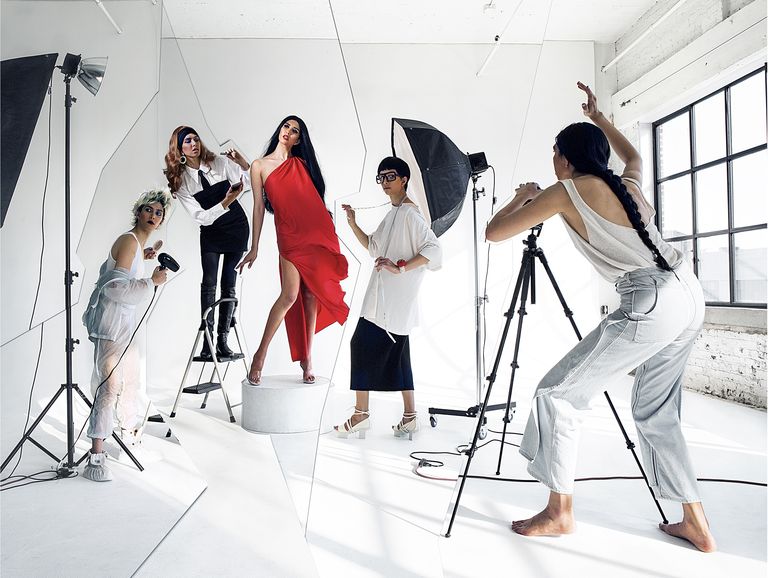 on the set of a photo shoot, a woman in a red dress is modeling as three individuals are around here directing her and working to fix her hair, makeup, and wardrobe, as another person shoots photos on a camera