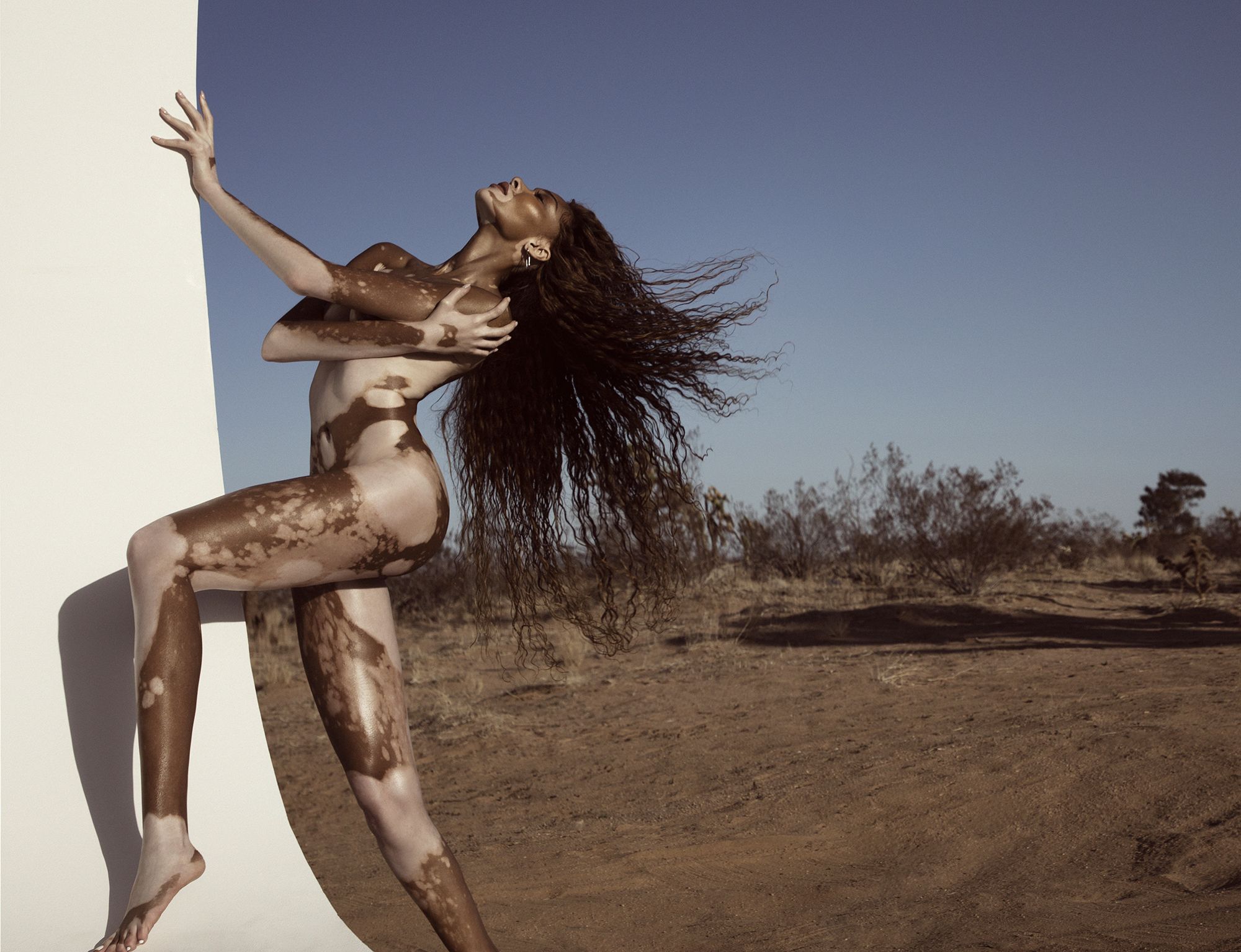 model winnie harlow standing sideways to the left, bare, head raised back towards sky, resting her left hand on white concrete wall with her right arm crossed under her left arm and right hand on left shoulder, covering her upper body, her left knee raised with left leg at 90 degrees in the air, on an open desert like landscape with blue sky