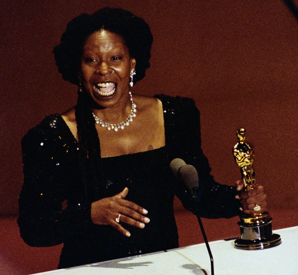 whoopi goldberg wears a black sparkly dress and holds an oscar as she accepts an award for best supporting actress, she is speaking into a microphone at a white podium with the statue in her left hand