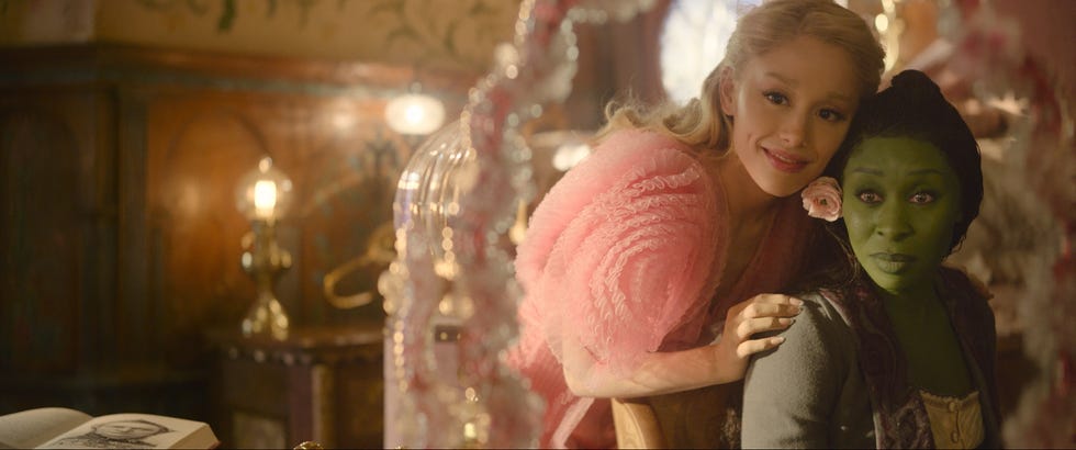 ariana grande and cynthia erivo on set of the movie wicked, erivo sits in profile and looks over her shoulder to the left as grande leans against her at the back of the chair