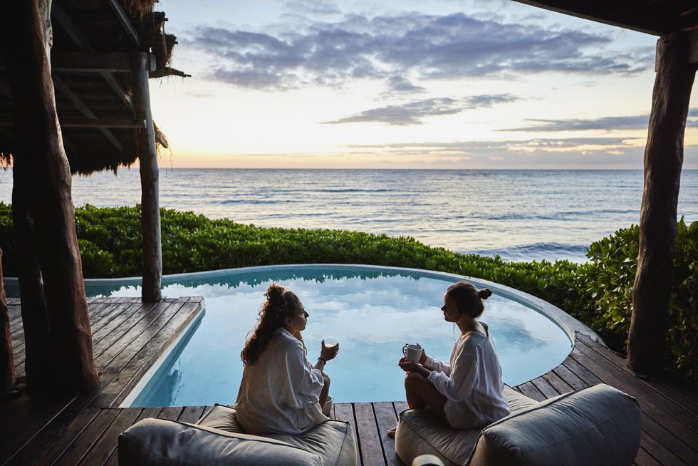 wide shot of female friends in discussion while sitting poolside at luxury suite of tropical resort at sunrise