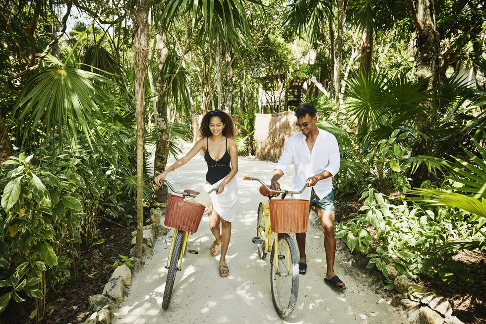 wide shot of smiling couple walking bikes on pathway in jungle at tropical resort
