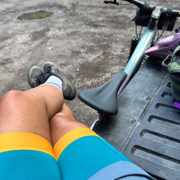 a person sits on a bench in bike shorts next to bike, helmet, backpack