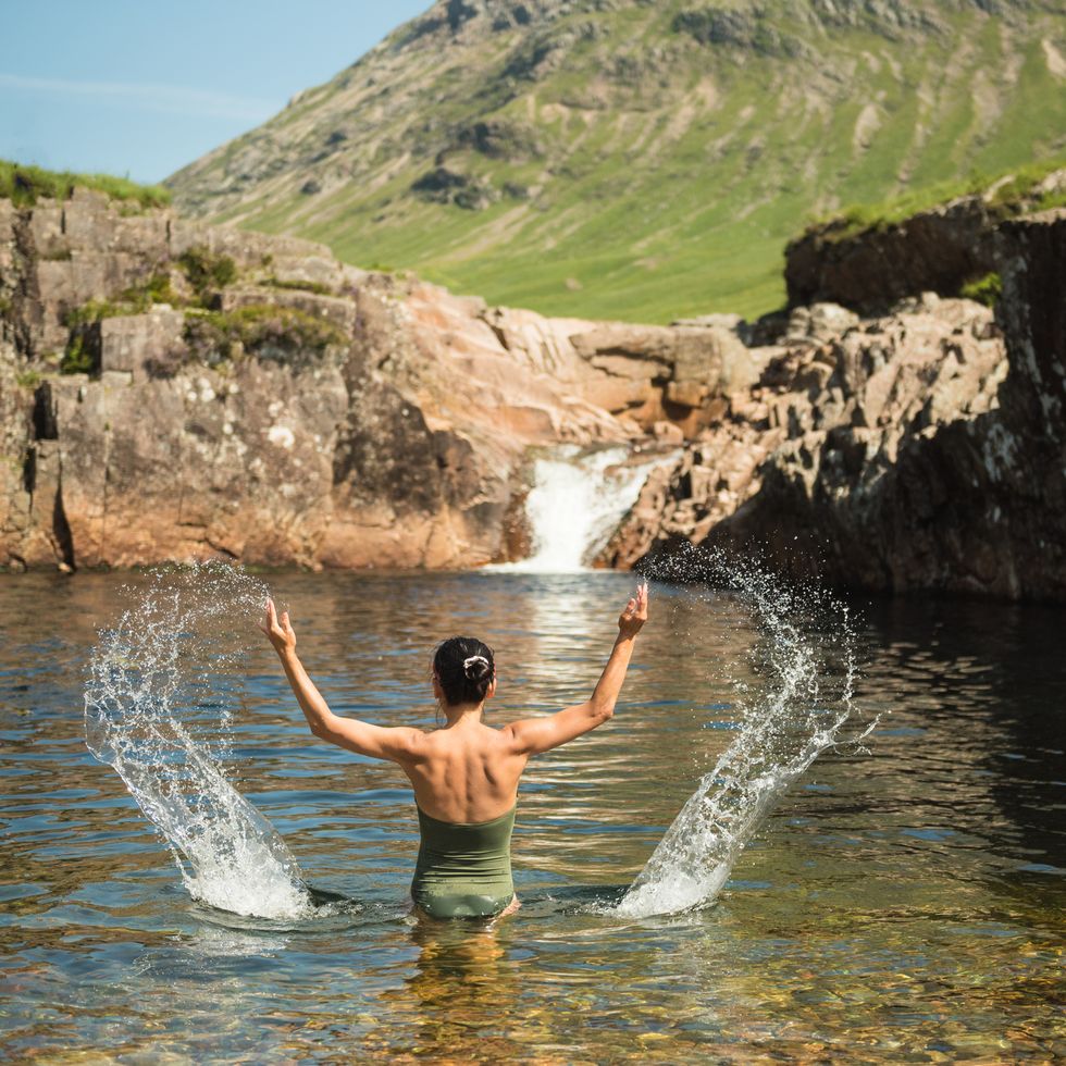 a woman wearing a green swimming suit splashes water with both hands while bathing in the river etive in glen etive, scottish highlands, uk, with a waterfall and a mountain in the background