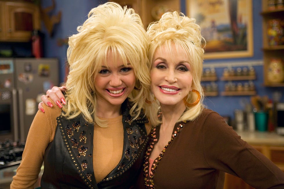 miley cyrus and dolly parton smile for a photo together and hug, cyrus wears a blonde wig styled to look like parton's hair