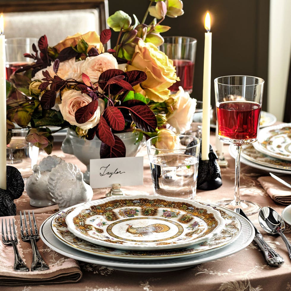 Thanksgiving Tablescape Ideas a beautifully arranged dining table featuring an assortment of plates, including a decorative one with a turkey design in the center the table is adorned with a centerpiece of soft colored roses and greenery elegant glassware includes a tall stem glass filled with red liquid and a clear glass of water a white candle stands lit next to a place card named 'taylor,' and small ornamental figures are also present on the table