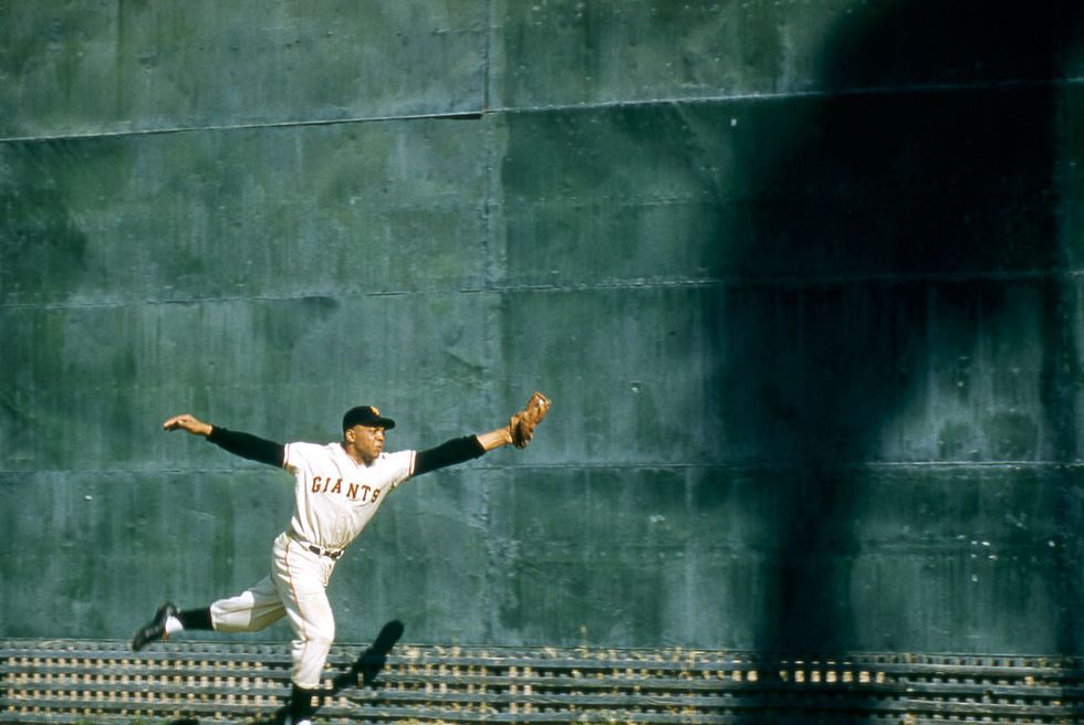 willie mays jumps up and reaches one hand with a baseball glove out to catch a baseball, he wears a giants baseball uniform and cap