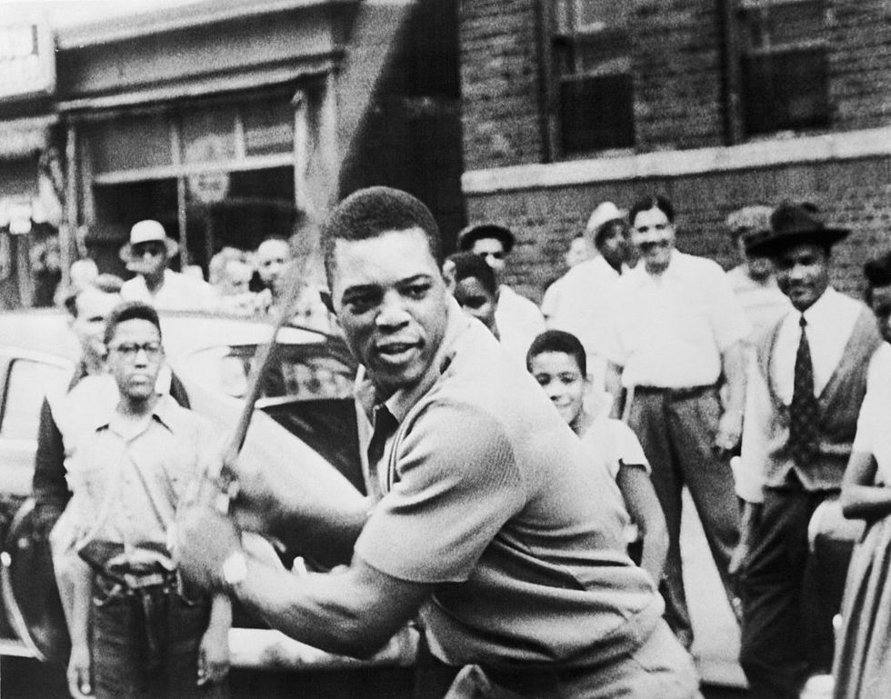 willie mays stands on a street and swings a stick as many children and adults watch behind him