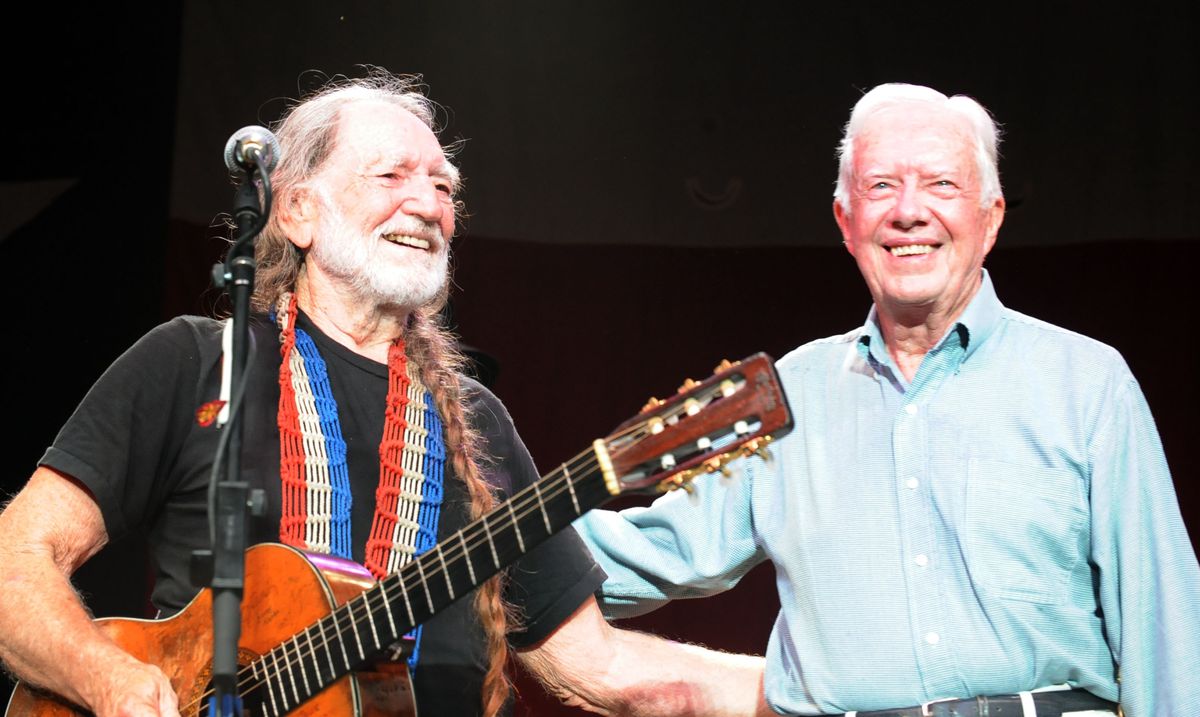 willie nelson holding a guitar and standing in front of a microphone while smiling and stnading next to a smiling jimmy carter