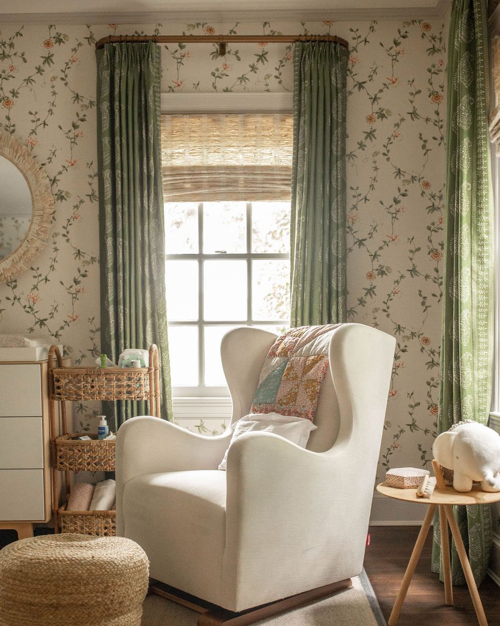 nursery, white rocking chair with pink and blue quilt, nude and grey area rug, wooden floor, floral wallpaper and green curtains