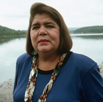 wilma mankiller poses for a photo while standing on a shore of a lake, she wears a blue shirt and long beaded necklace