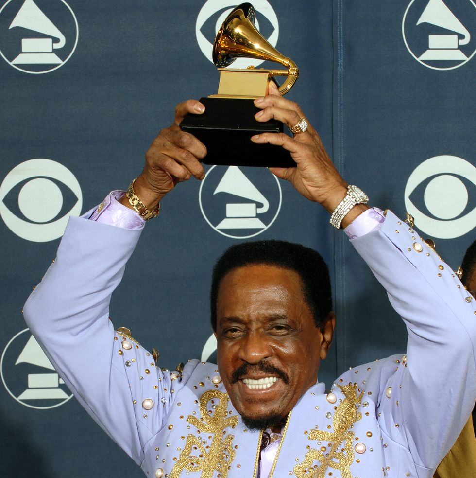 ike turner holds a grammy award above his head and smiles, he is wearing a lavendar suit jacket with pearl and gold studs and gold accents