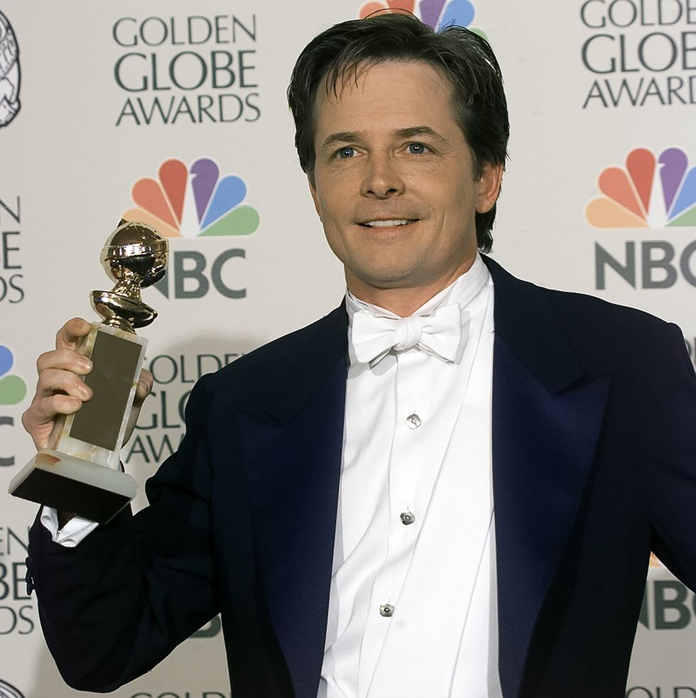 michael j fox holds a golden globe trophy in his right hand and smiles as he looks past the camera, he is wearing a black suit jacket, white shirt and white bow tie and stands in front of a golden globe awards backdrop
