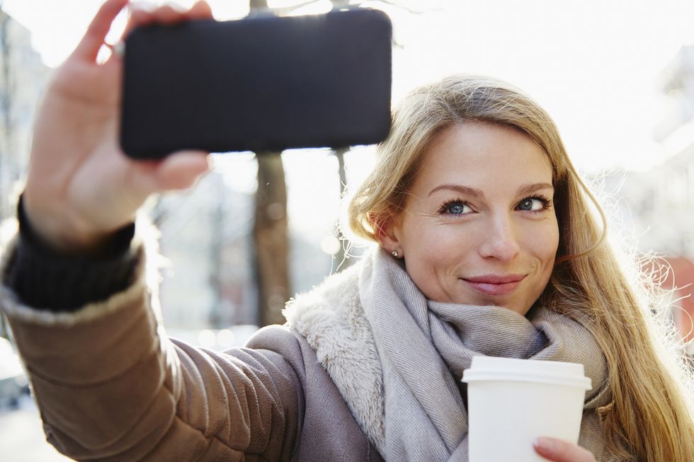 handsome woman walking the city streets in winter taking a selfie and holding a coffee to go, backlit